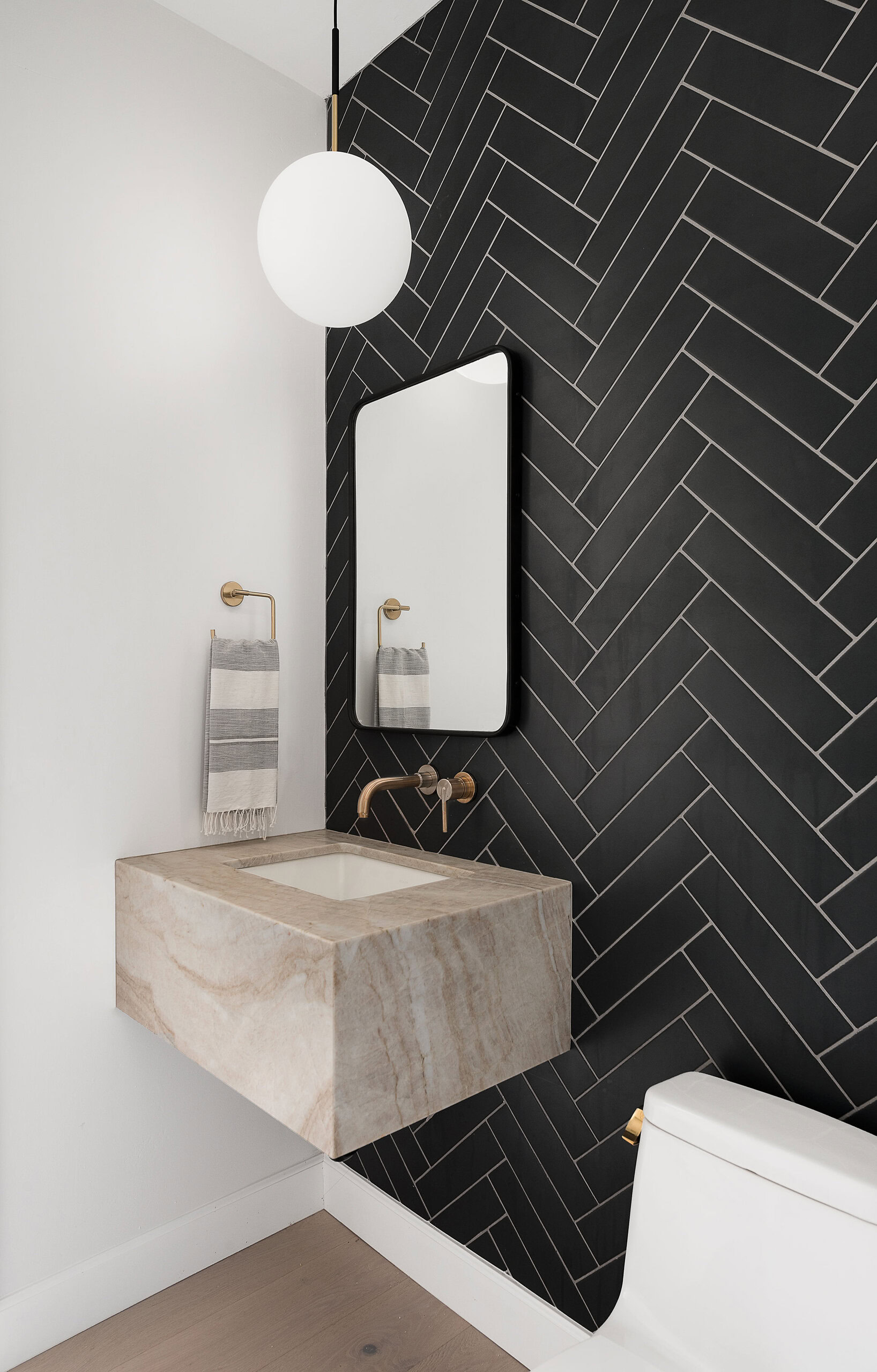 Black herringbone tile wall in bathroom with beige stone vanity and gold fixtures.