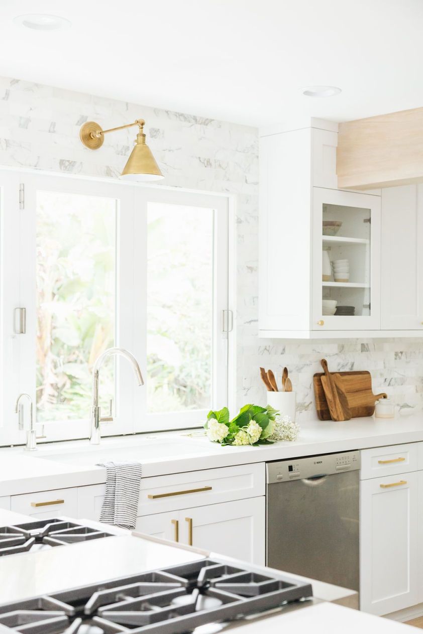 Bright white kitchen featuring marble backsplash, gold fixtures, and natural light from large window.