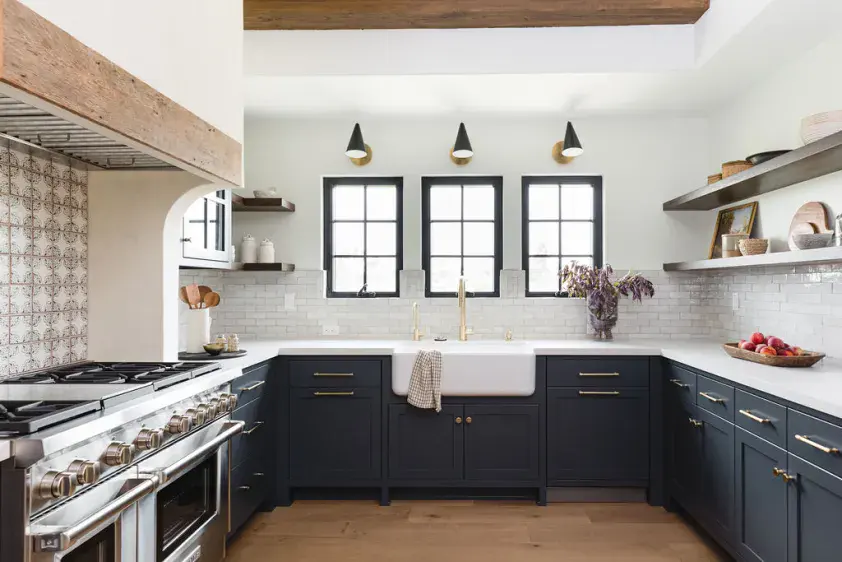 Dark blue kitchen cabinets with white farmhouse sink and open shelving, featuring modern black windo.
