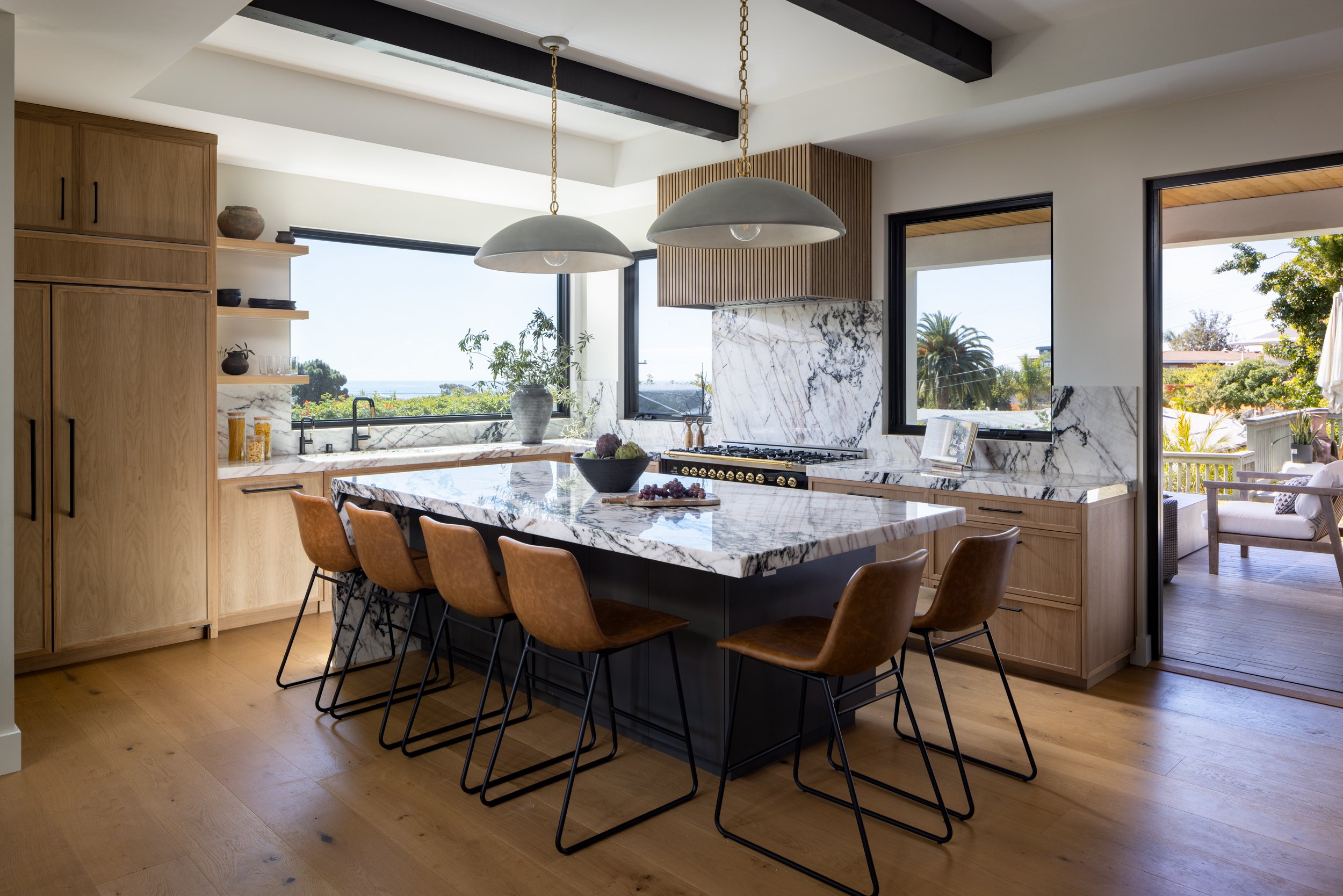 Modern kitchen with marble island, wooden cabinets, and large windows overlooking outdoor scenery.