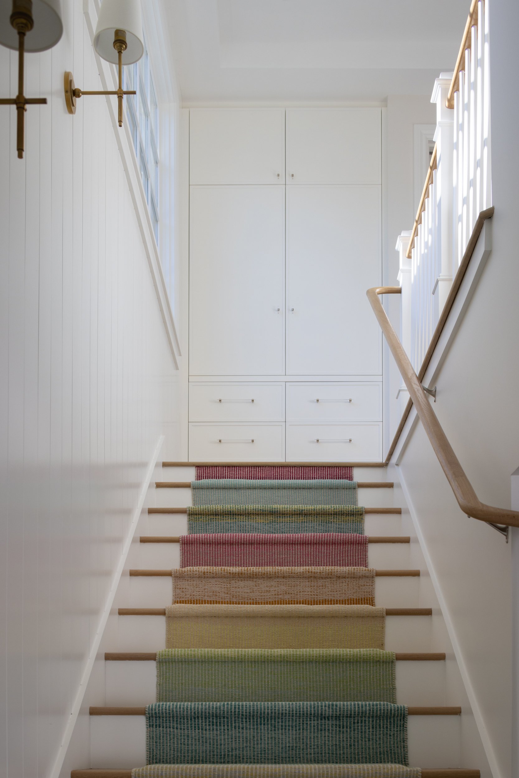 Bright white staircase with colorful striped runner and wooden handrail.