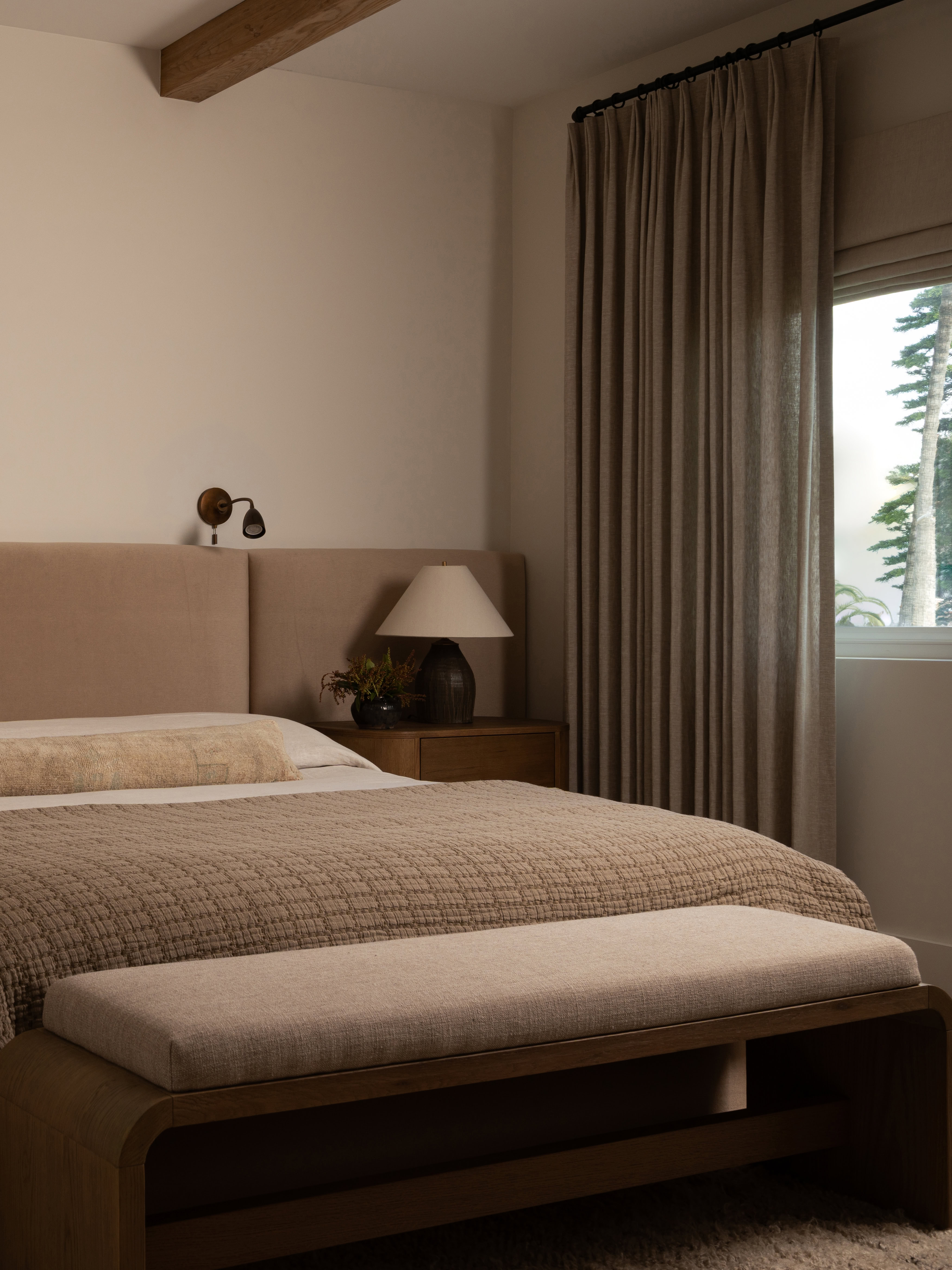 Modern bedroom with beige bedding, bedside lamp, and window view of trees.