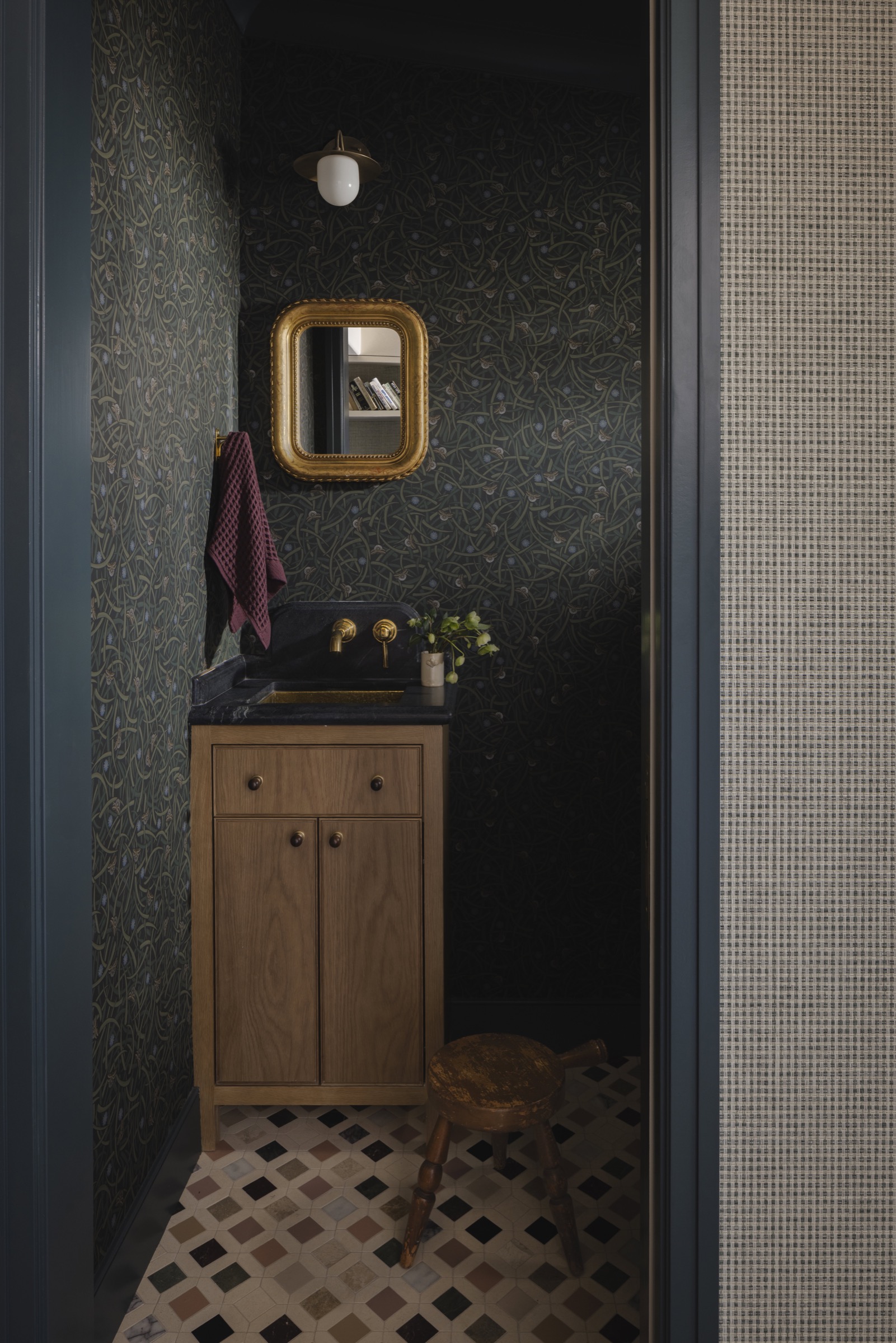 Small bathroom with dark wallpaper, wooden vanity, gold mirror, and vintage stool.
