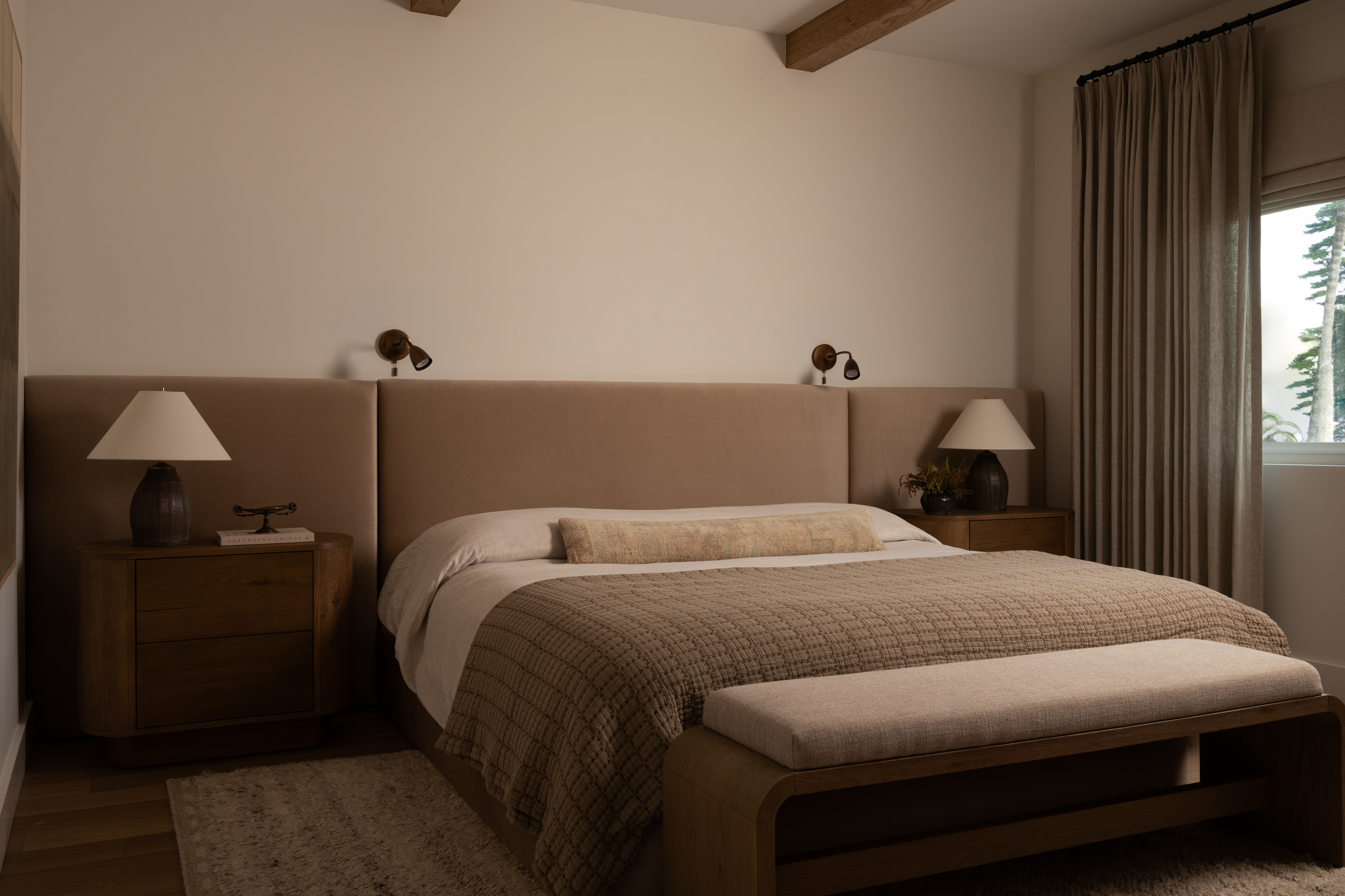 Modern bedroom with beige headboard, wooden nightstands, and natural light from window.