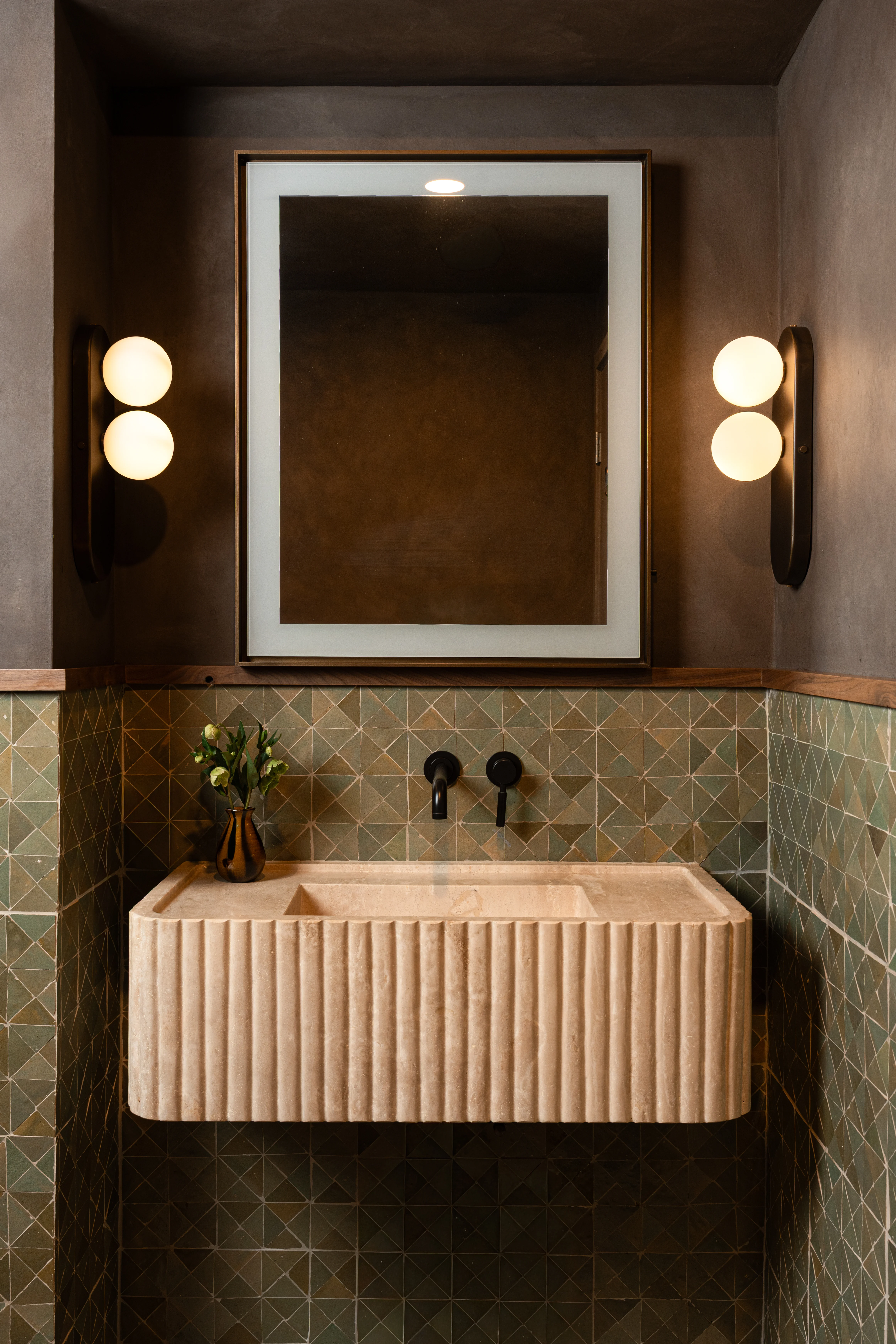 Moody powder room with stone tile, fluted vanity, and round sconces.