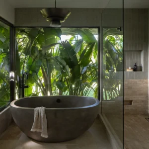 Stone soaking tub set into a glass corner surrounded by tropical foliage.