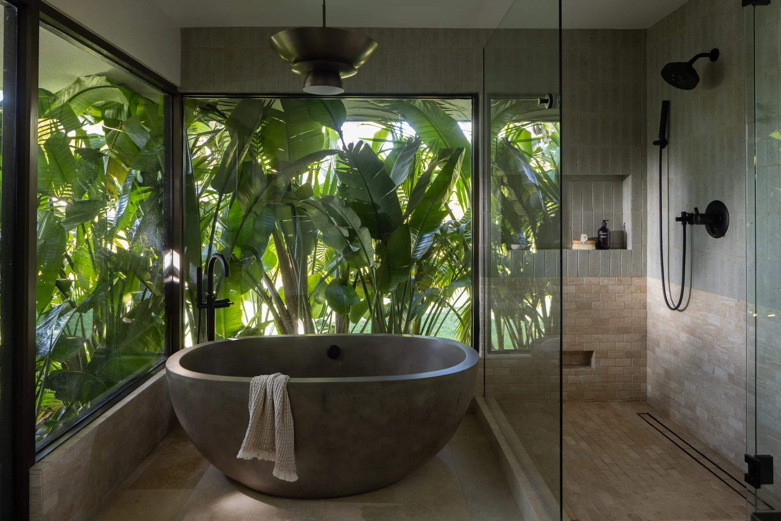 Stone soaking tub set into a glass corner surrounded by tropical foliage.