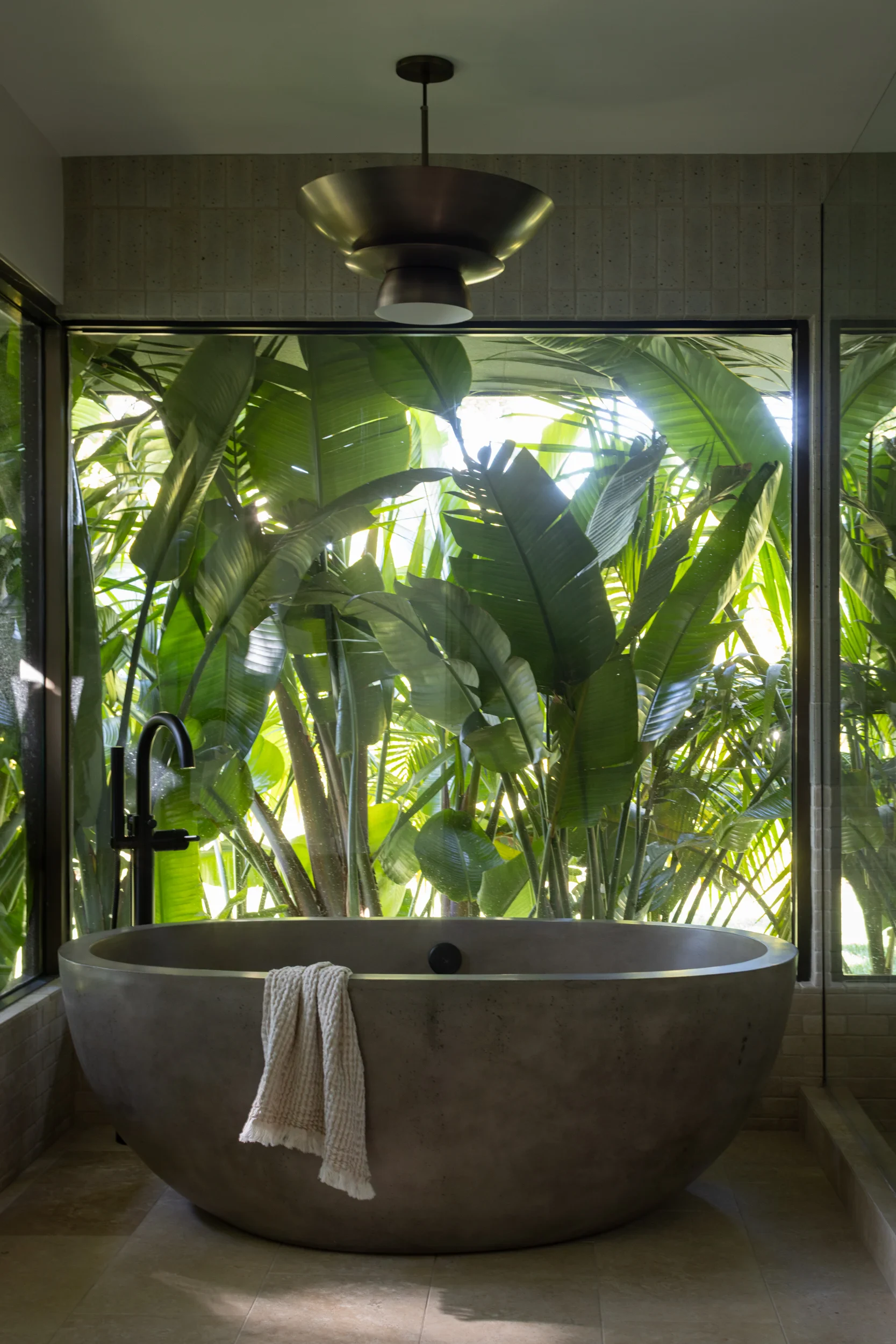 Freestanding stone tub beneath a bronze pendant with banana-leaf garden views.